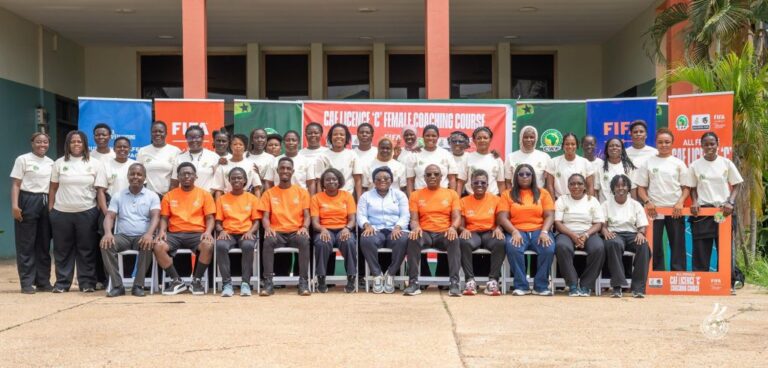 Group of men and women in orange and white shirts posing for a photo in front of a FIFA coaching course banner outdoors.