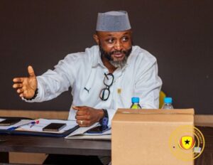 Bearded man in a light shirt and gray kufi speaks at a table, gesturing with his left hand amid papers and two water bottles on the desk.