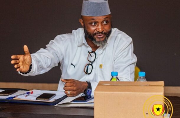 Bearded man in a light shirt and gray kufi speaks at a table, gesturing with his left hand amid papers and two water bottles on the desk.