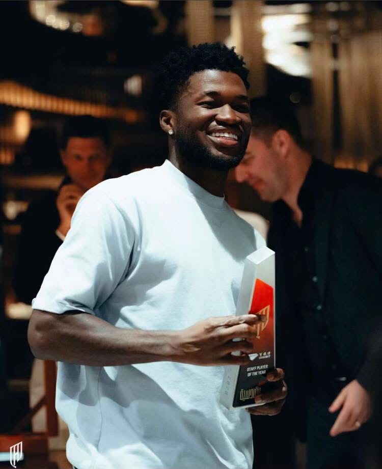 Smiling Black man in a light blue shirt holds a tall award plaque with red design in a dim venue, with people blurred in the background.