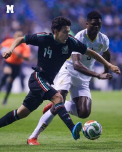 Two soccer players chase the ball during a match, one in a dark Mexico kit (#19) and the other in white, on a green field with a ball at their feet.