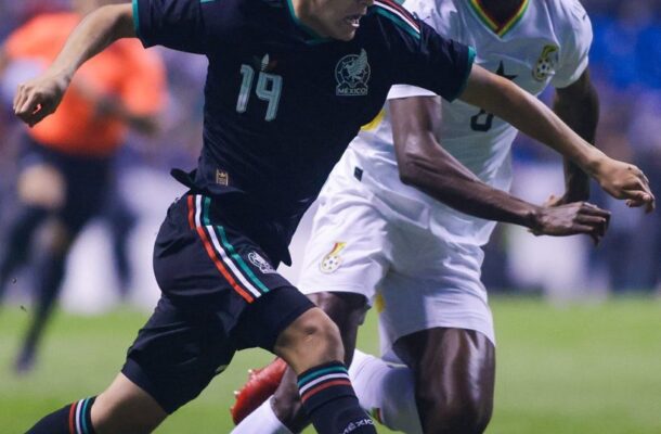 Two soccer players chase the ball during a match, one in a dark Mexico kit (#19) and the other in white, on a green field with a ball at their feet.