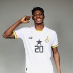 Smiling Black man in a white soccer jersey with number 20, pointing to his head, against a gray background.