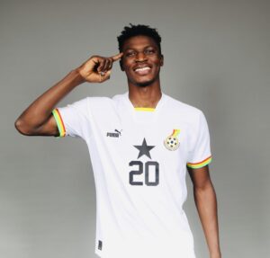 Smiling Black man in a white soccer jersey with number 20, pointing to his head, against a gray background.
