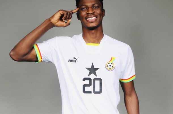 IMG_0643 | Ghanaguardian.com Smiling Black man in a white soccer jersey with number 20, pointing to his head, against a gray background.