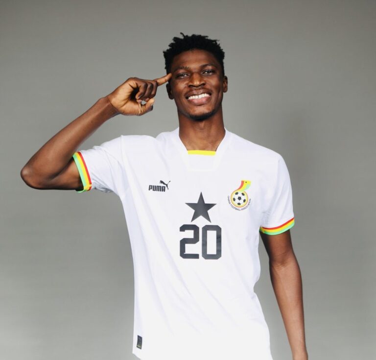 Smiling Black man in a white soccer jersey with number 20, pointing to his head, against a gray background.
