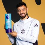 Male footballer in a Manchester United jersey holds a blue Premier League Player of the Season trophy and smiles at the camera.