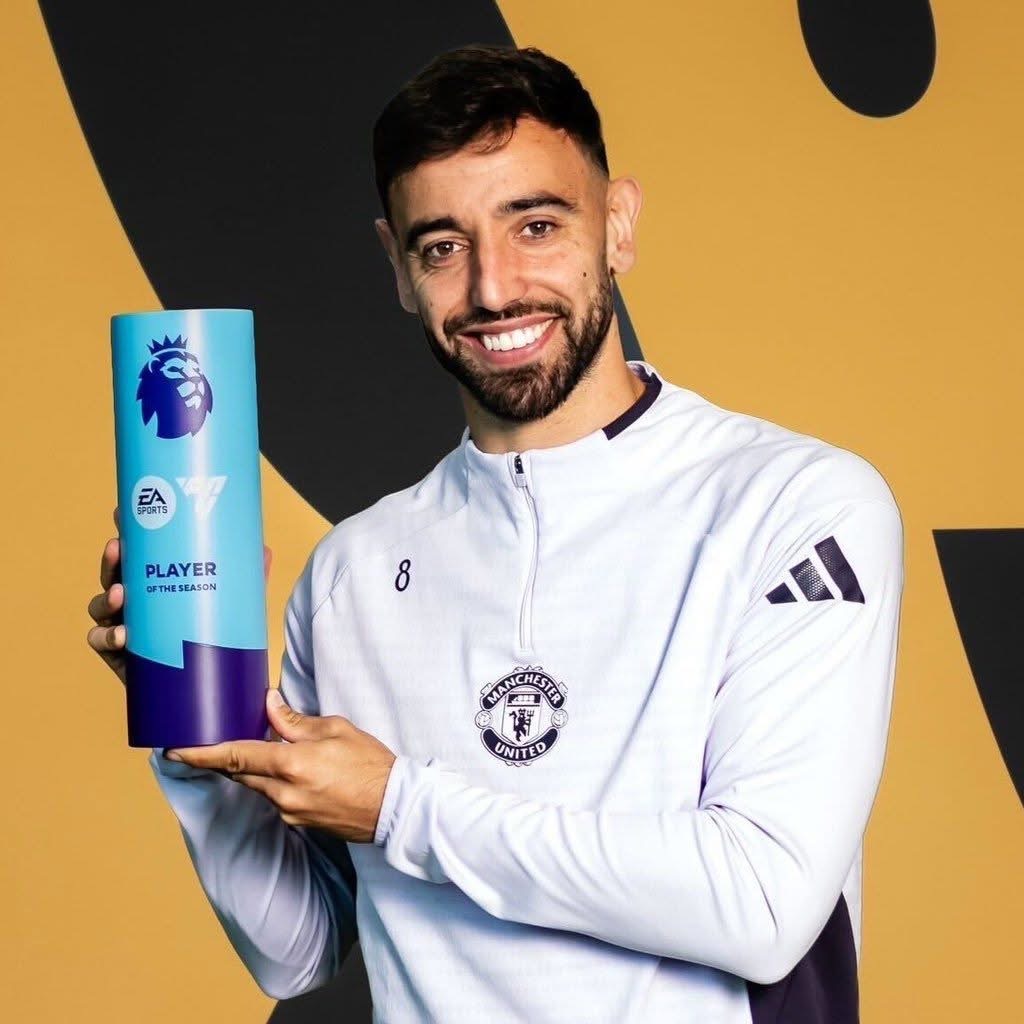 Male footballer in a Manchester United jersey holds a blue Premier League Player of the Season trophy and smiles at the camera.