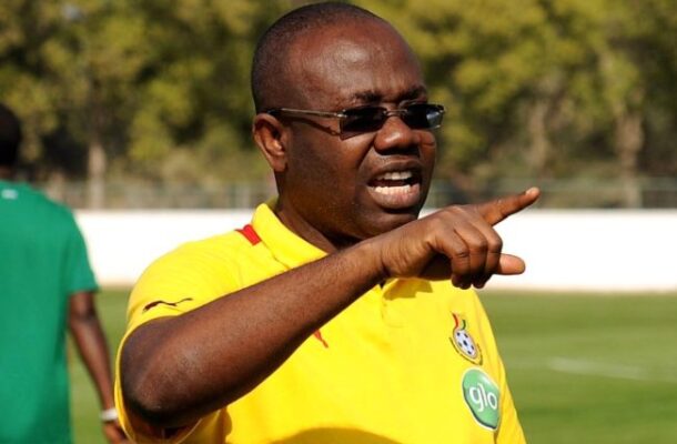 Dark-skinned man in a yellow soccer jersey and sunglasses points and speaks on a sunny field.