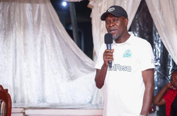 Male presenter wearing a white polo with a 'HardPesa' logo and a black cap speaks into a handheld microphone under a draped white backdrop on stage.