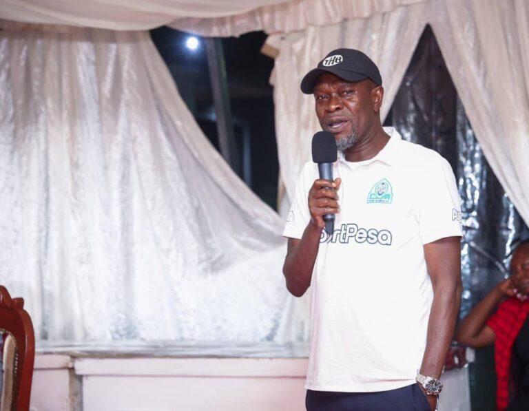 Male presenter wearing a white polo with a 'HardPesa' logo and a black cap speaks into a handheld microphone under a draped white backdrop on stage.