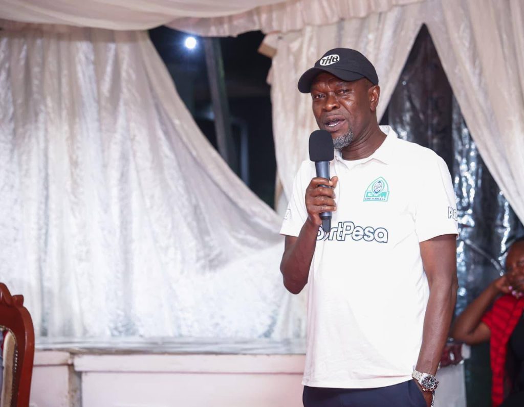 Male presenter wearing a white polo with a 'HardPesa' logo and a black cap speaks into a handheld microphone under a draped white backdrop on stage.