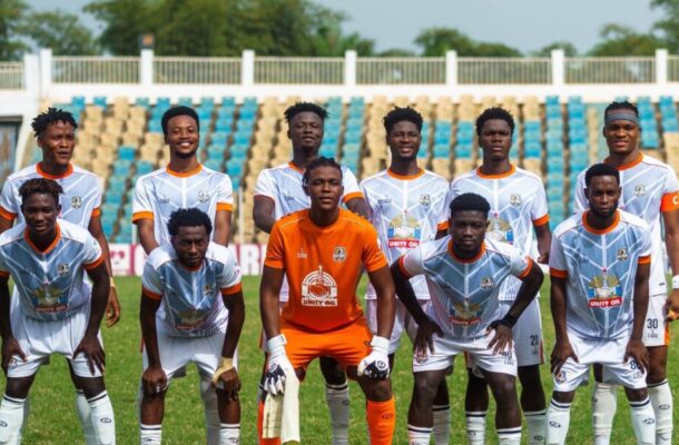 Soccer team posing for a group photo on a sunny field; goalkeeper in orange kit stands center, teammates in white jerseys with orange trim behind and in front, stadium seating in the background.