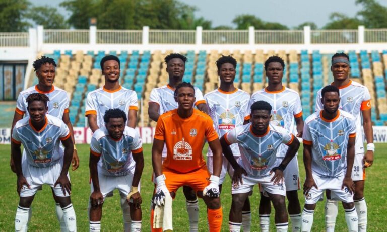 Soccer team posing for a group photo on a sunny field; goalkeeper in orange kit stands center, teammates in white jerseys with orange trim behind and in front, stadium seating in the background.