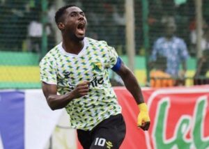 Soccer player in a green-yellow patterned jersey celebrates with mouth open and fist raised on the field.