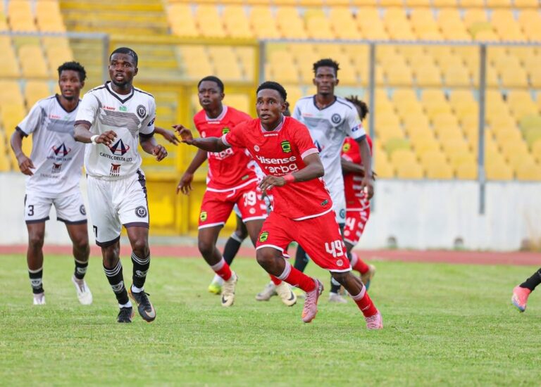 Soccer players chase the ball on a grass field during a match, with yellow stadium seats in the background, under bright light far in the stands.