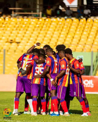 Football team in purple, red, and yellow kits huddles on the field before a match.