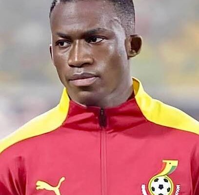 Male soccer player wearing a red and yellow training jacket with a Ghana Football Association crest on the chest, looking off to the side