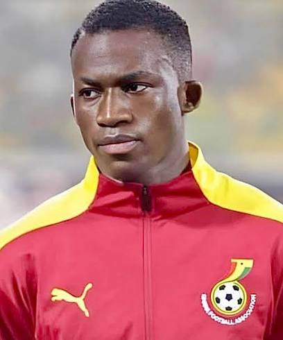 Male soccer player wearing a red and yellow training jacket with a Ghana Football Association crest on the chest, looking off to the side