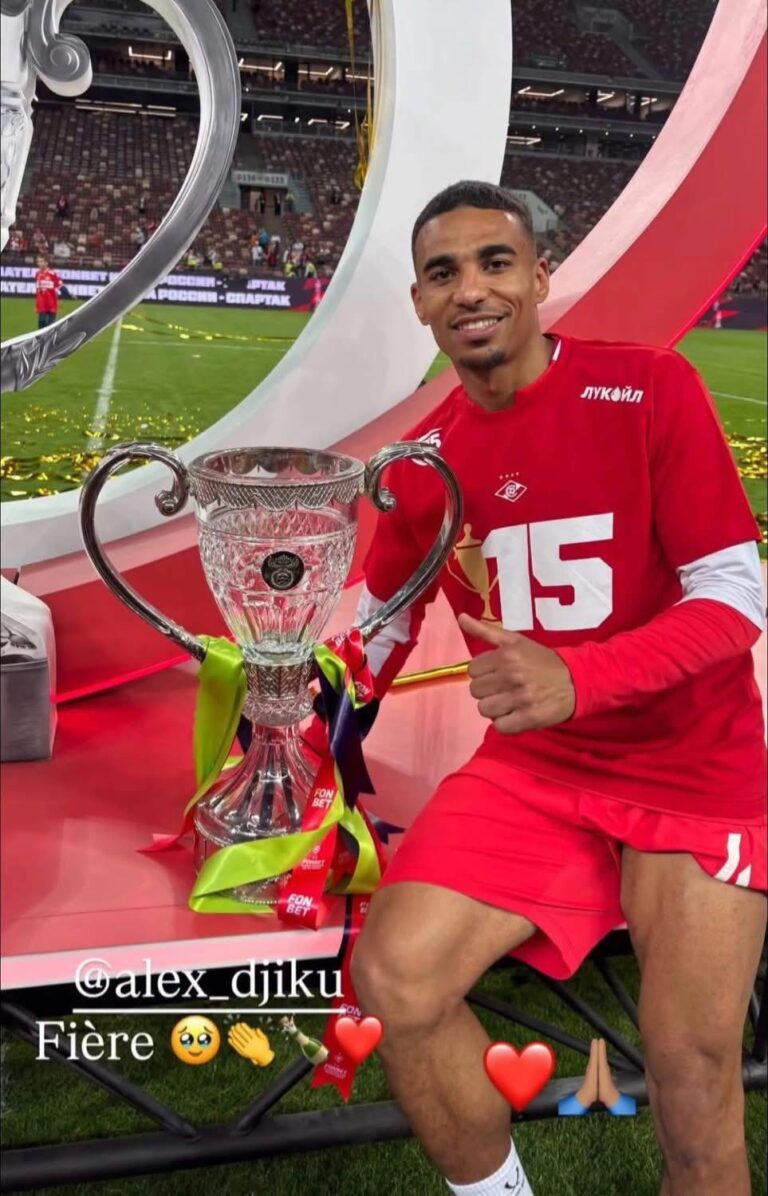 Footballer in red jersey sits beside a large crystal trophy on a red podium, giving a thumbs-up with a stadium background.