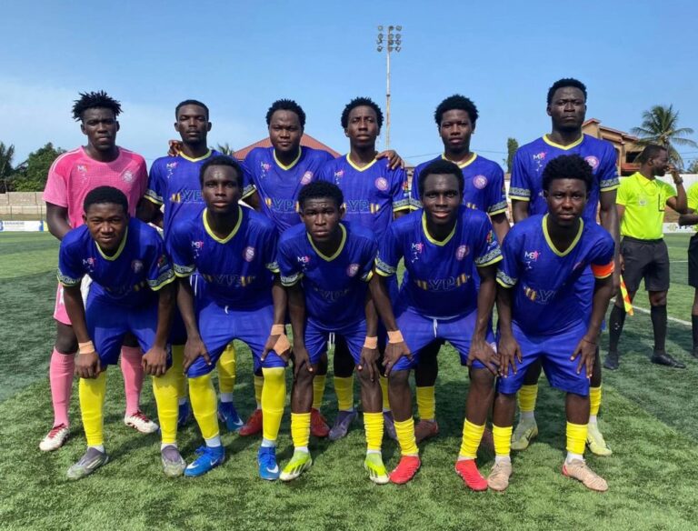 Group of 11 young soccer players in blue uniforms with yellow socks posing for a team photo on a sunny grass field.