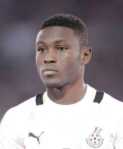 Close-up of a male soccer player in a white jersey featuring a team crest on the chest, neutral expression.