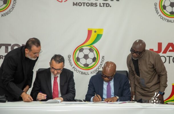 Four men in suits signing documents at a table during a formal agreement, with a Ghana Football Association banner in the background.
