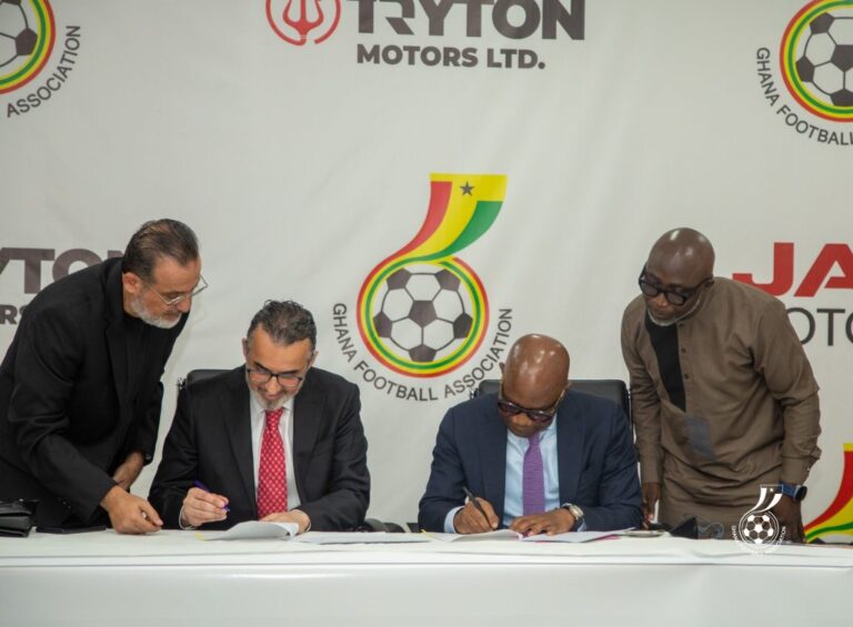 Four men in suits signing documents at a table during a formal agreement, with a Ghana Football Association banner in the background.