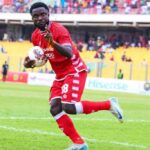 Soccer player in a red kit sprinting on the green field with the ball, yellow-stands crowd in the background