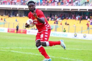 Soccer player in a red kit sprinting on the green field with the ball, yellow-stands crowd in the background