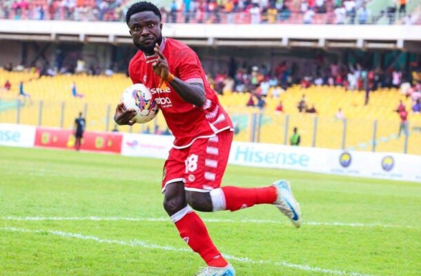 Soccer player in a red kit sprinting on the green field with the ball, yellow-stands crowd in the background