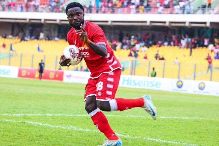 Soccer player in a red kit sprinting on the green field with the ball, yellow-stands crowd in the background