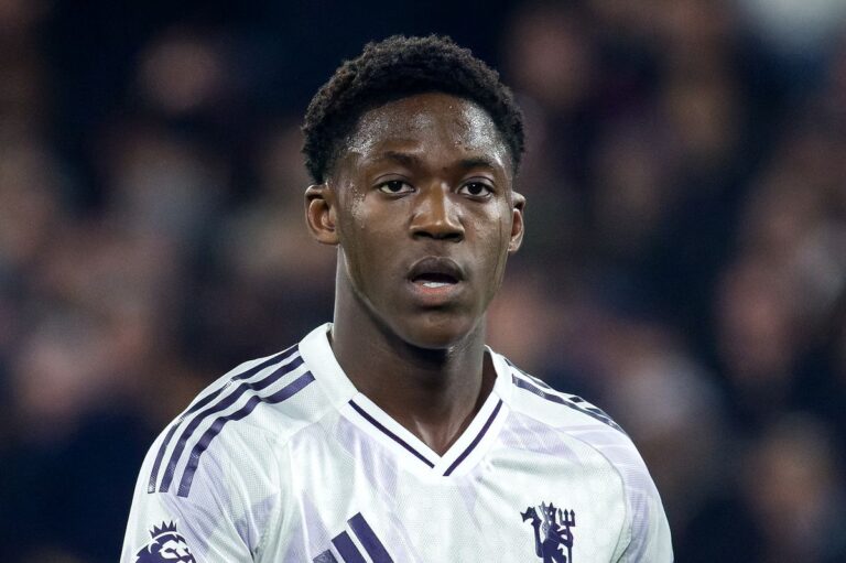 Soccer player in a white jersey with purple stripes, head-and-shoulders shot on the field, looking ahead.