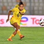 Female soccer player in a yellow uniform lunges to kick the ball on a green field during a match.