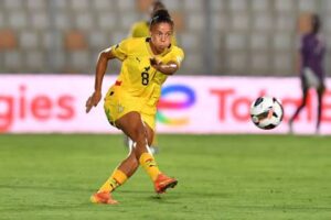Female soccer player in a yellow uniform lunges to kick the ball on a green field during a match.