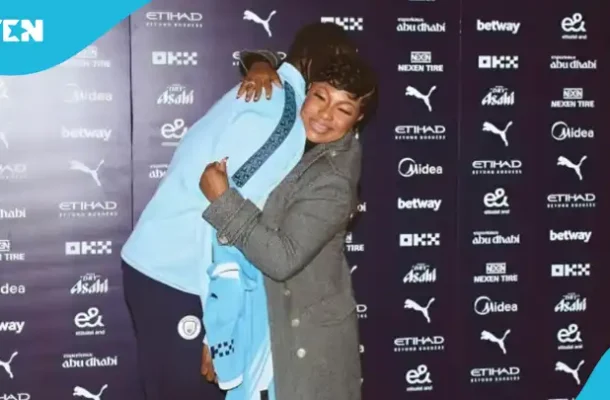 IMG_0746 | Ghanaguardian.com Two people hug on a blue-carpet backdrop filled with sponsor logos, one in a gray coat and the other in a light blue jacket, sharing a warm moment at an event.