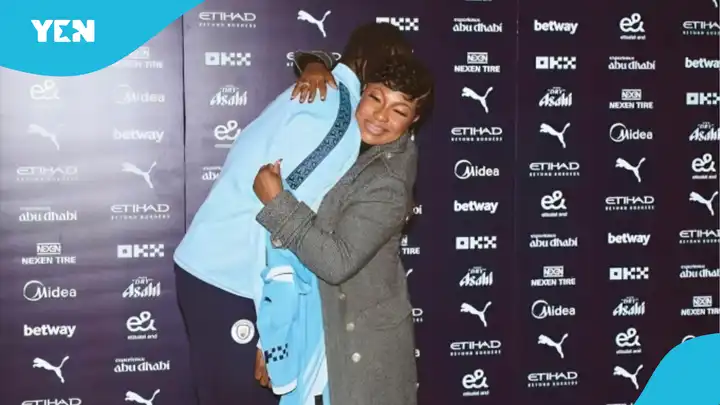 Two people hug on a blue-carpet backdrop filled with sponsor logos, one in a gray coat and the other in a light blue jacket, sharing a warm moment at an event.