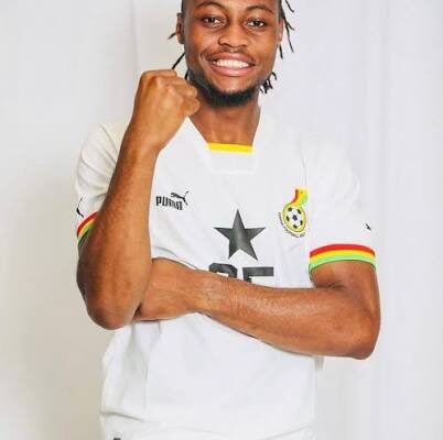 Footballer in a white jersey with a star emblem celebrates, fist raised and a smile on a light backdrop.