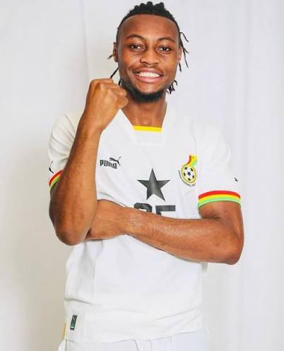 Footballer in a white jersey with a star emblem celebrates, fist raised and a smile on a light backdrop.