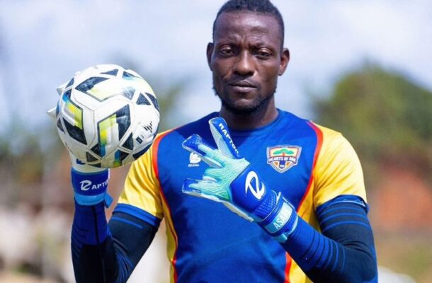 Goalkeeper in blue-yellow kit juggling a soccer ball, wearing blue gloves, focused on the ball outdoors