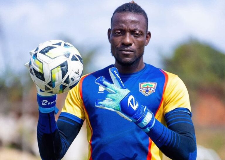 Goalkeeper in blue-yellow kit juggling a soccer ball, wearing blue gloves, focused on the ball outdoors