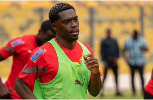 Soccer player in a red jersey and neon green training bib running on a pitch with yellow seats in the background.