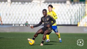 Soccer player in black kit dribbles a yellow ball as a yellow-clad defender pursues in a stadium field.