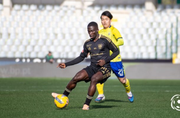 Soccer player in black kit dribbles a yellow ball as a yellow-clad defender pursues in a stadium field.
