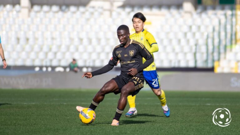 Soccer player in black kit dribbles a yellow ball as a yellow-clad defender pursues in a stadium field.