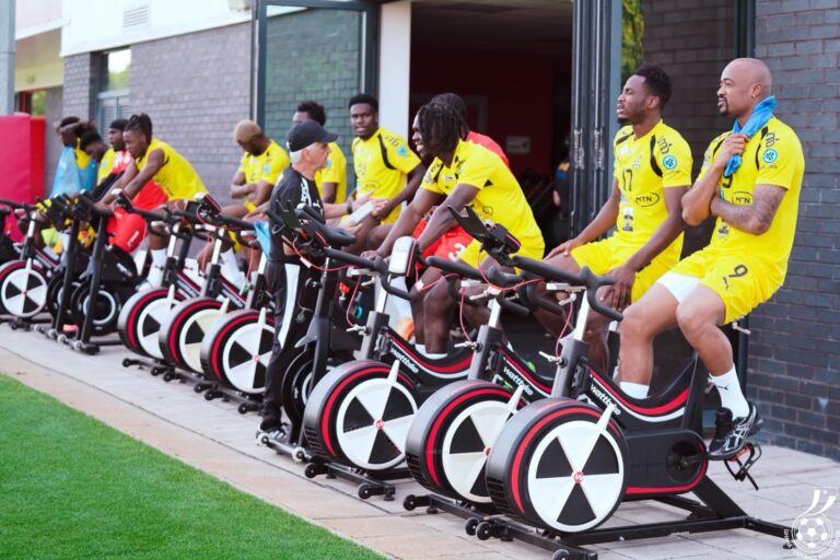 Group of athletes in yellow uniforms sit on black and red stationary bikes lined up outside a building outside a gym entrance.