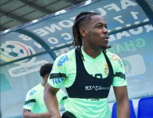 Soccer player with dreadlocks wearing neon green training kit and a black bib, standing near the dugout with another player in the background.