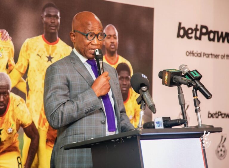 Man in a gray plaid suit speaks into a microphone at a press conference with a football-themed backdrop behind him.