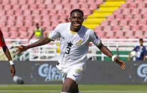 Soccer player in a white jersey (#3) celebrating with arms outstretched on a green pitch with stadium seats behind him, during a match.