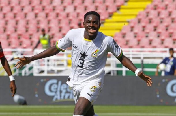 Soccer player in a white jersey (#3) celebrating with arms outstretched on a green pitch with stadium seats behind him, during a match.